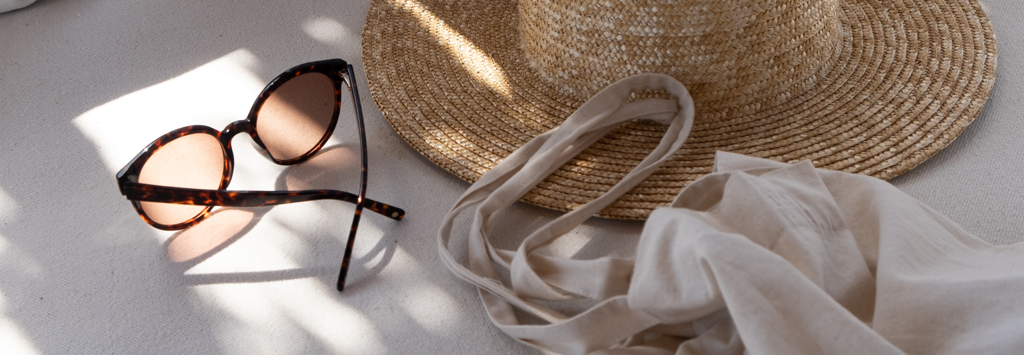 A lifestyle scene with tortoiseshell sunglasses, a woven straw hat, and a beige linen cloth placed on a neutral-toned surface. Natural sunlight creates soft shadows, evoking a summery, relaxed vibe.