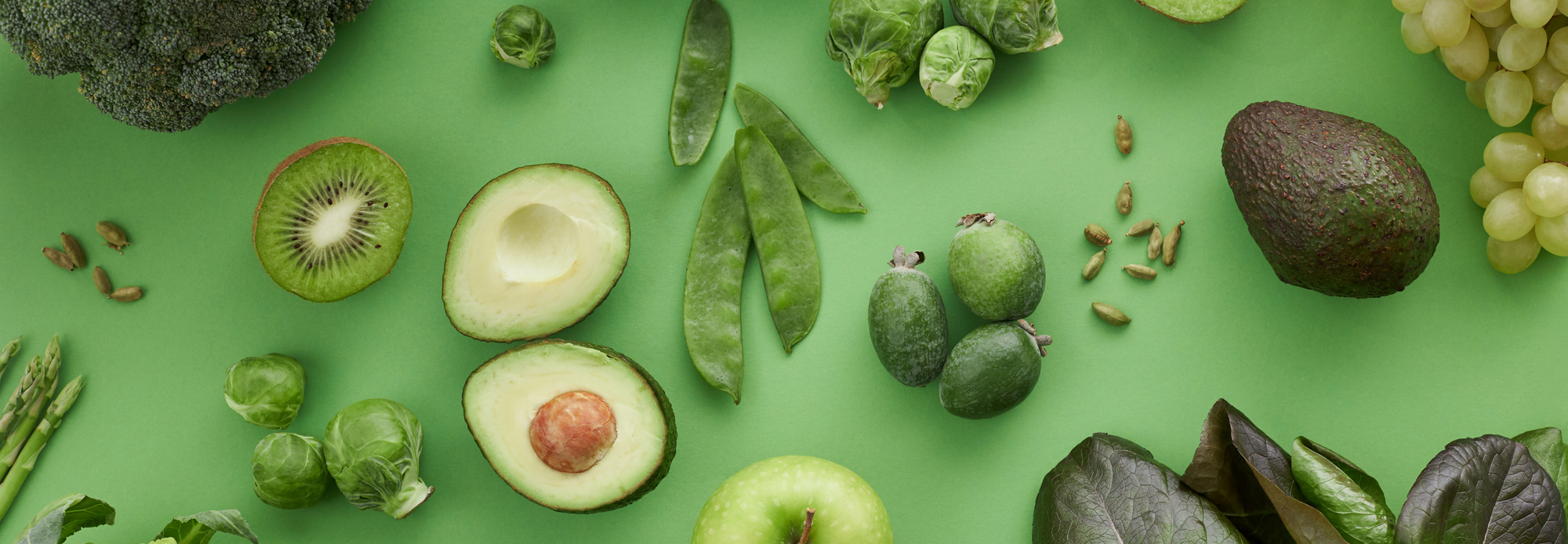 A flat lay of green fruits and vegetables, including avocado, kiwi, Brussels sprouts, green apple, and snow peas, arranged on a bright green background. The composition emphasizes freshness and healthy ingredients.