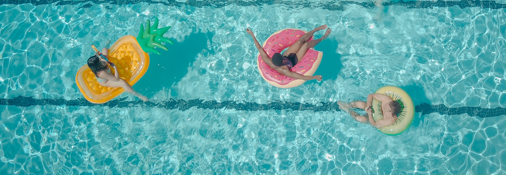 A top-down view of three people relaxing on colorful inflatable floats in a swimming pool. The clear water and playful floats give a vibrant, summery feel.