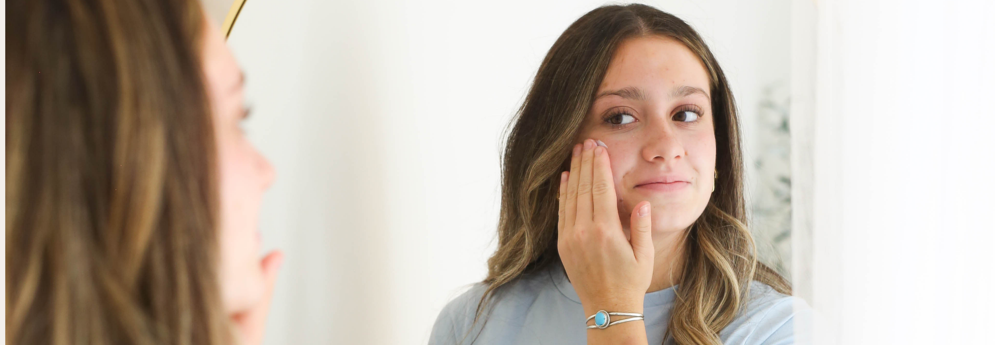A young woman looks at herself in a mirror while gently applying skincare product to her cheek. She wears a light blue top and a silver bracelet.