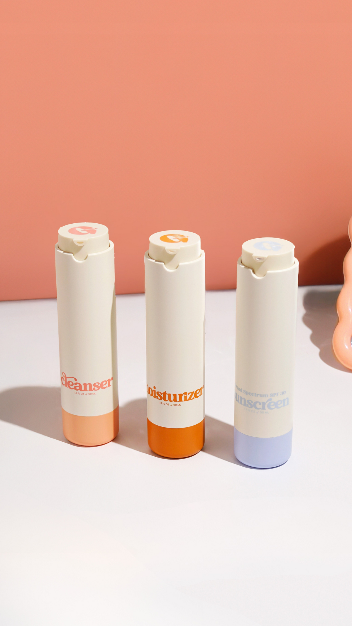Three minimalist Girlyish skincare bottles labeled cleanser, moisturizer, and sunscreen, standing in a row on a peach and white background