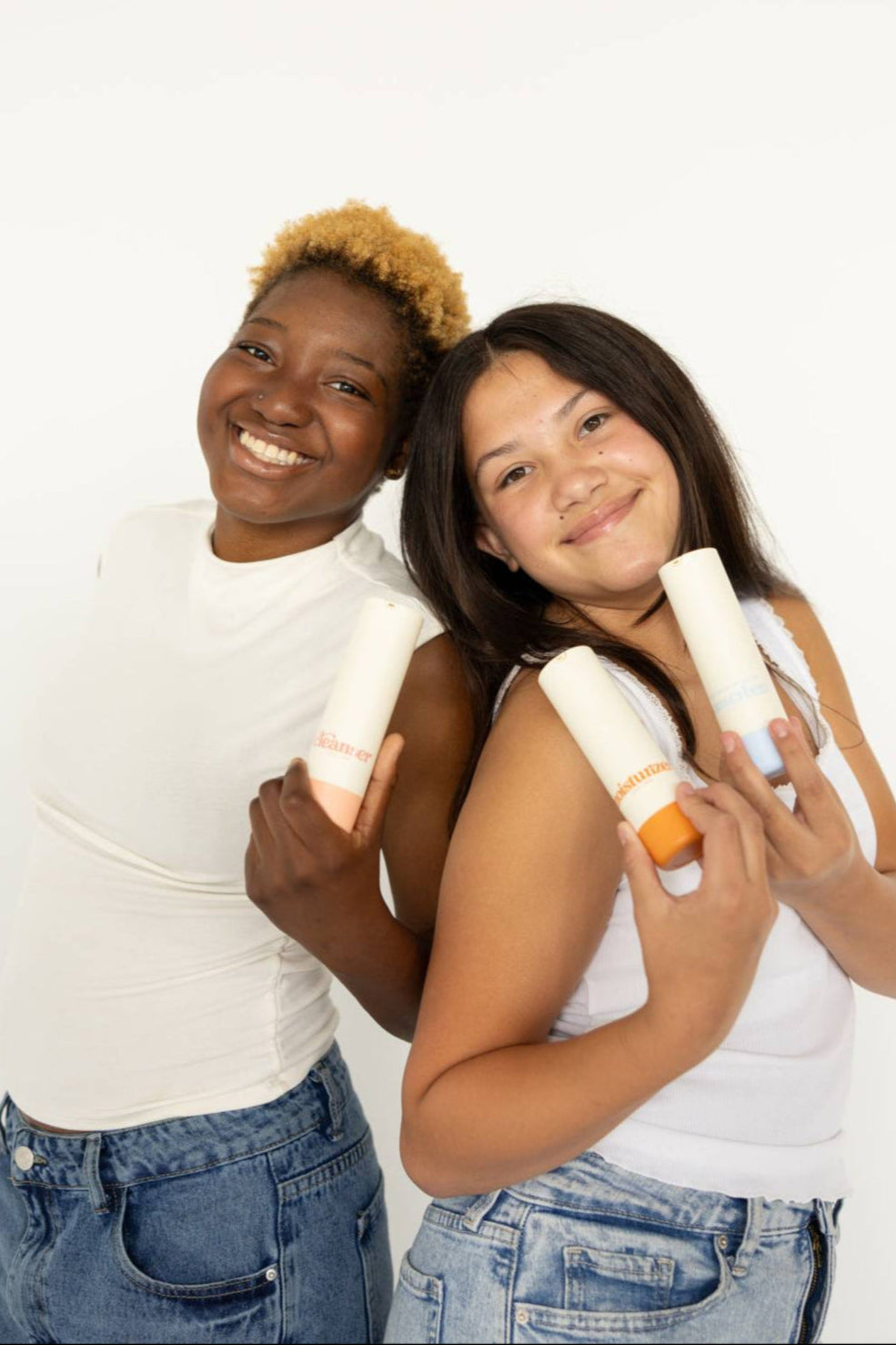 Two smiling girls in white tops holding Girlyish Skincare cleanser, moisturizer, and sunscreen bottles against a plain white background