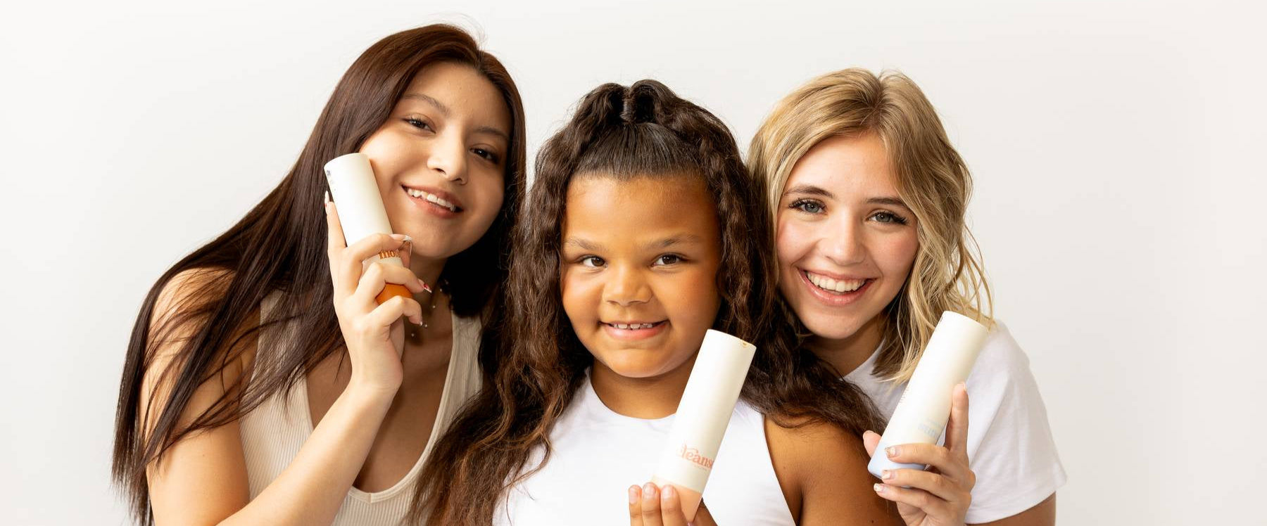 Three young girls smiling in white tops holding Girlyish Skincare bottles in front of a white background