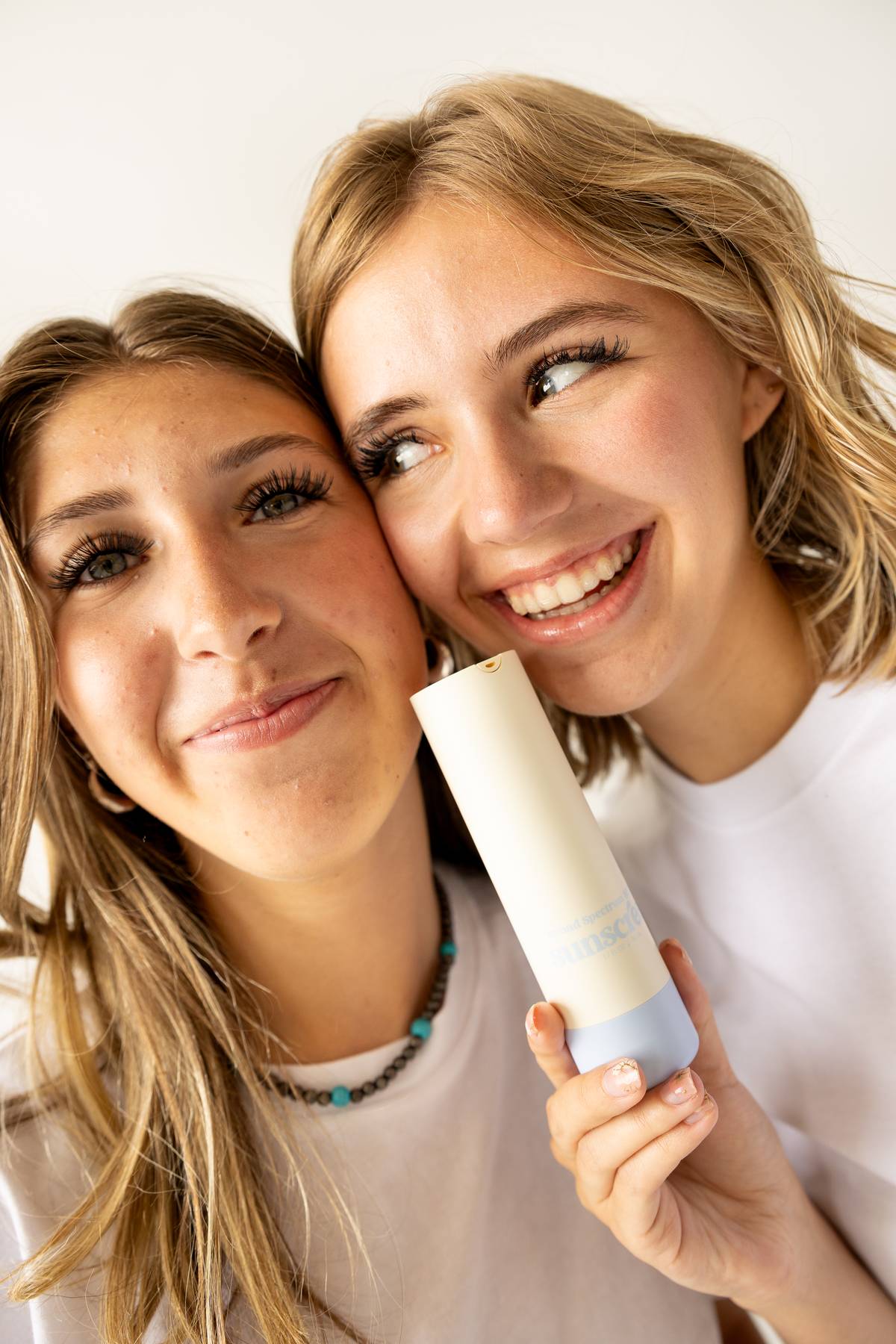 Two smiling girls pose closely together, one holding a skincare bottle labeled "sunscreen." Both are wearing white tops, and their faces are brightly lit, showcasing their clear skin and joyful expressions.