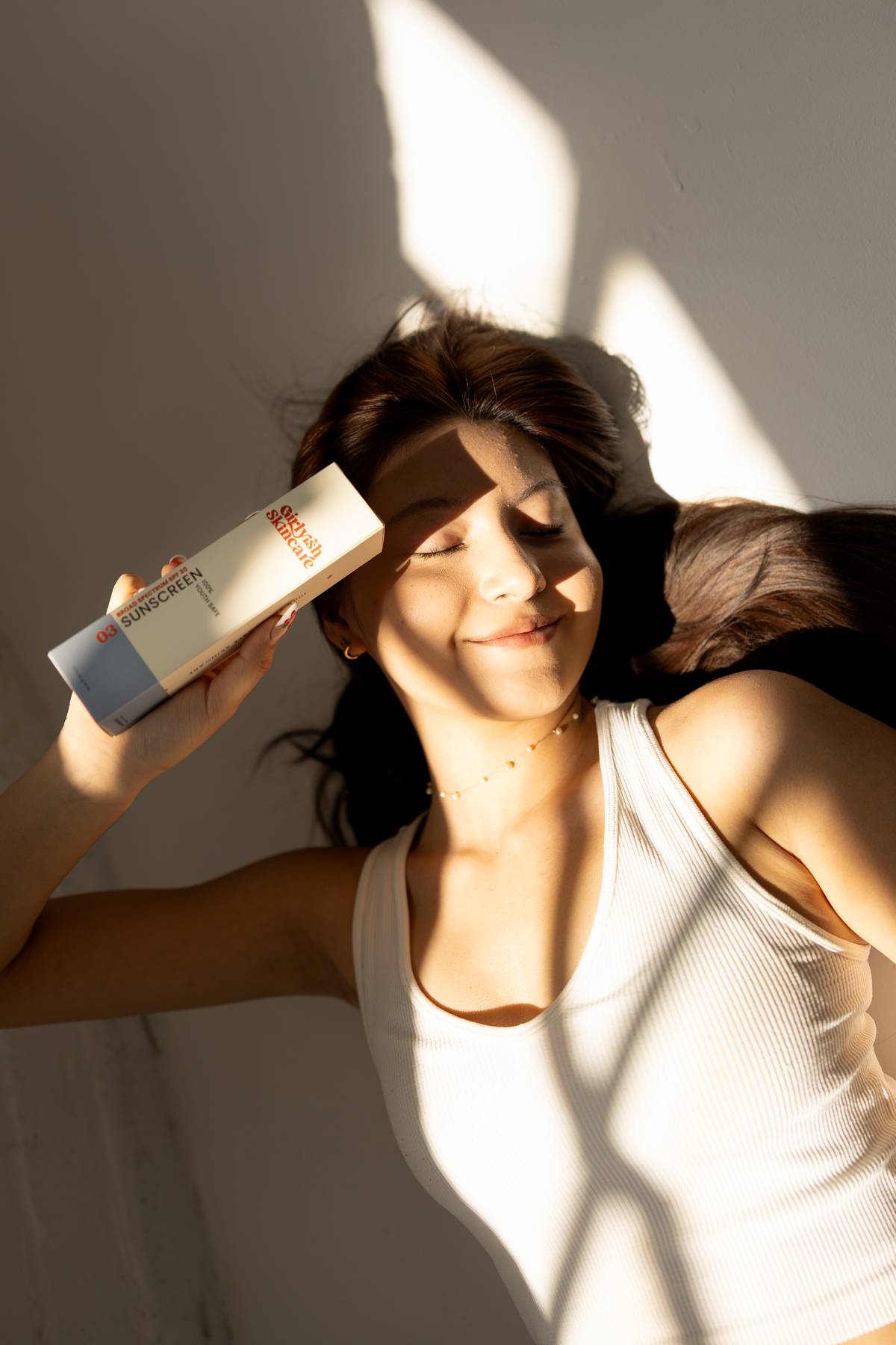 A teenage girl with long hair lies in warm sunlight against a white wall, eyes closed and smiling gently while holding a box labeled "Sunscreen" near their forehead. They are wearing a white tank top, and soft shadows fall across the photo.