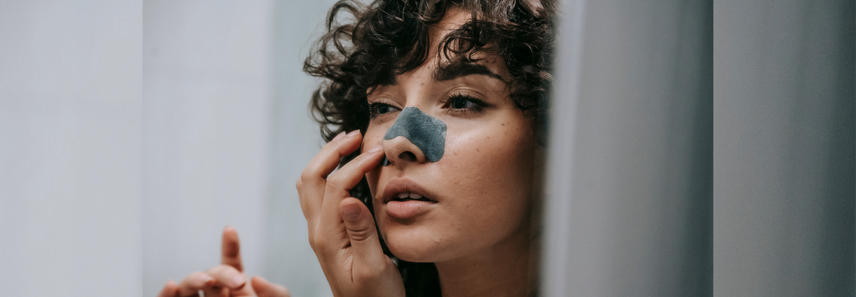 A person with curly hair applies a nose pore strip while looking into a mirror. The image captures a close-up of their face, framed between vertical edges of a bathroom mirror.