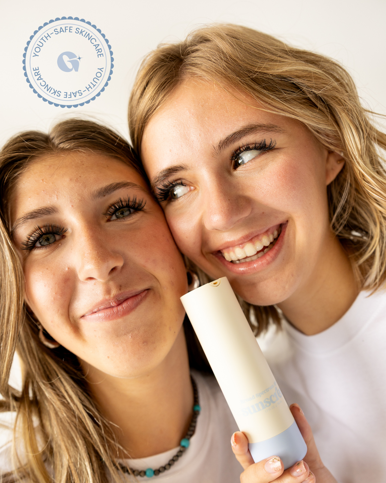 Two smiling girls pose closely together, one holding a skincare bottle labeled "sunscreen." Both are wearing white tops, and their faces are brightly lit, showcasing their clear skin and joyful expressions.