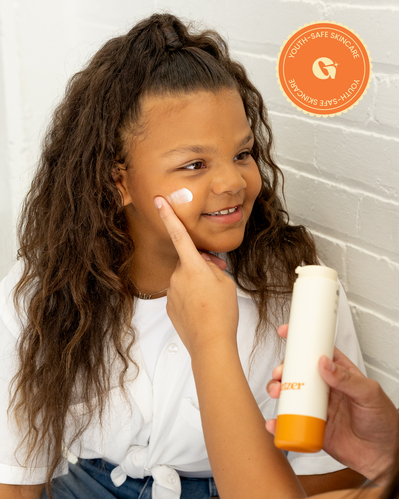 A young girl with long curly hair sits against a white brick wall, smiling as another girl applies moisturizer to her cheek. The second person holds a bottle labeled "moisturizer," and both are dressed casually in light-colored tops and jeans.