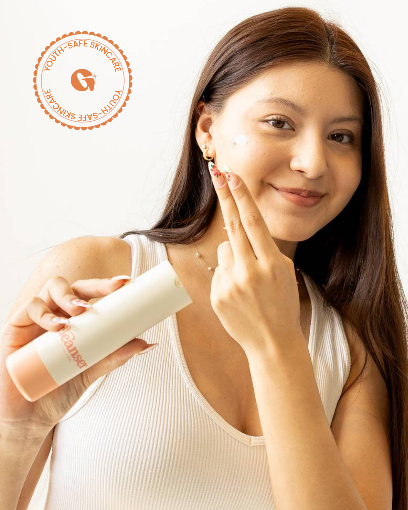 A young woman with long dark hair smiles while applying a dab of skincare product to her cheek. She is holding a bottle labeled “cleanser” and wearing a white top and blue jeans, standing against a light background.