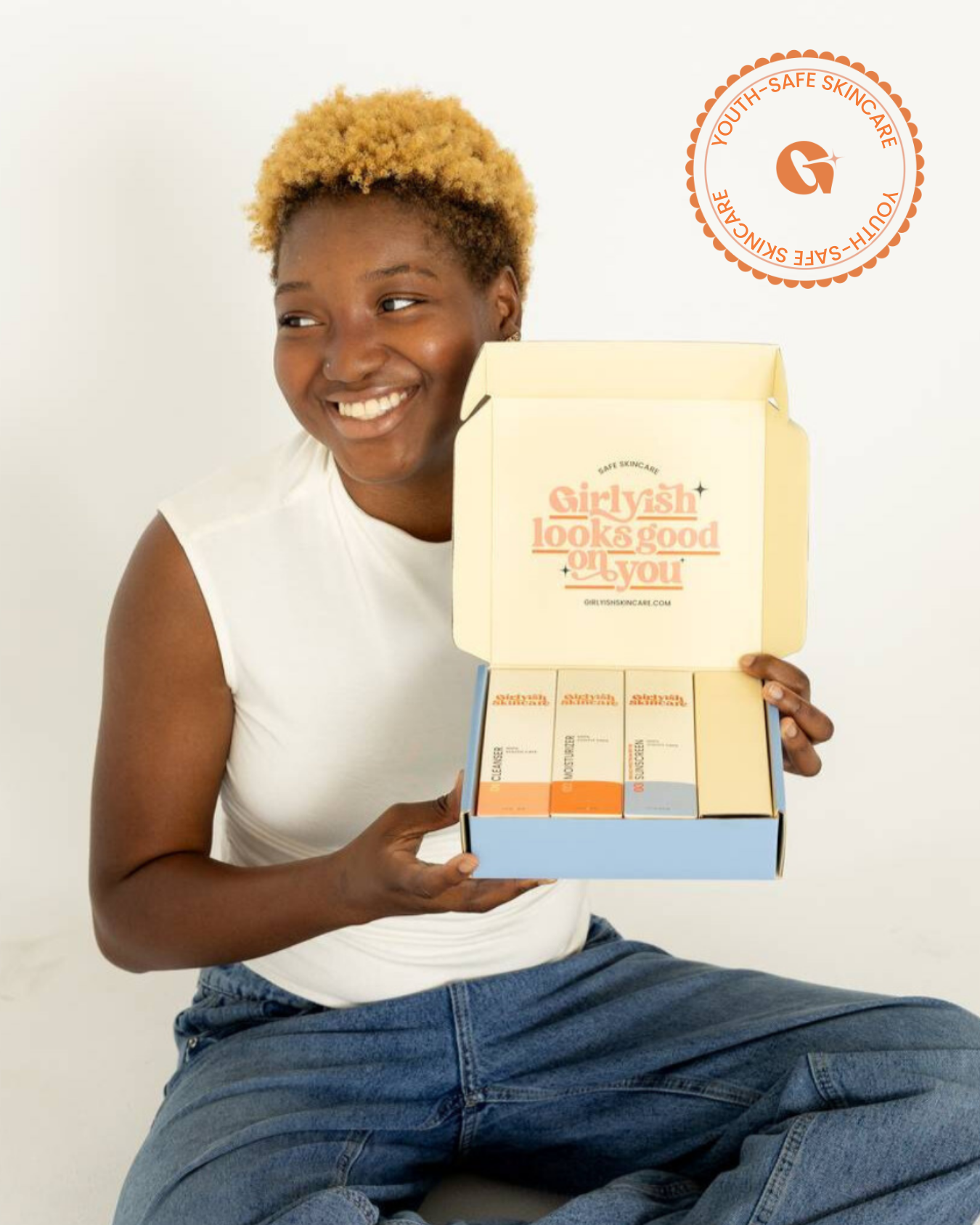 A smiling girl wearing a white sleeveless top and blue jeans, sits cross-legged on the floor holding an open skincare product box. The inside of the box lid reads "Girlyish looks good on you" with several colorful product bottles neatly arranged inside.