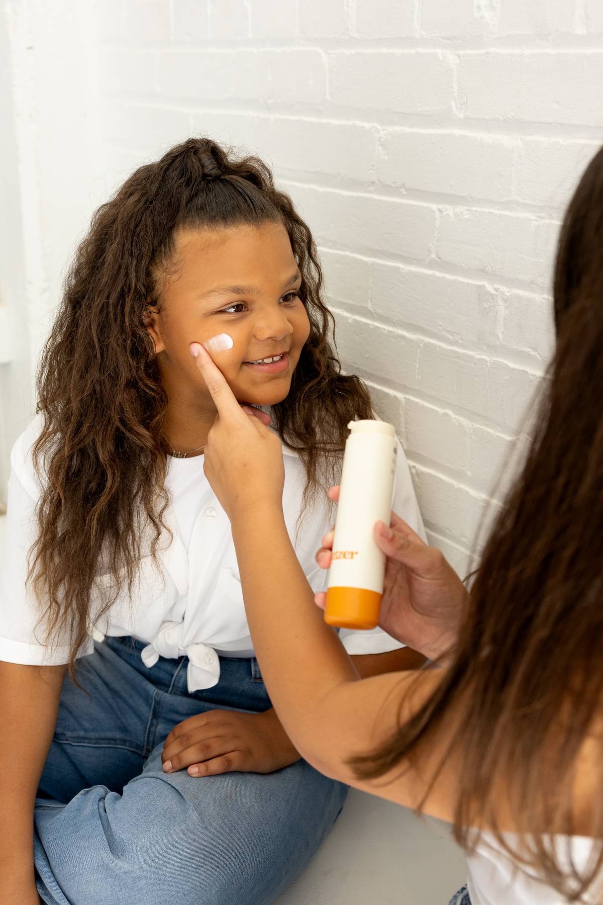 A young girl with long curly hair sits against a white brick wall, smiling as another girl applies moisturizer to her cheek. The second person holds a bottle labeled "moisturizer," and both are dressed casually in light-colored tops and jeans.