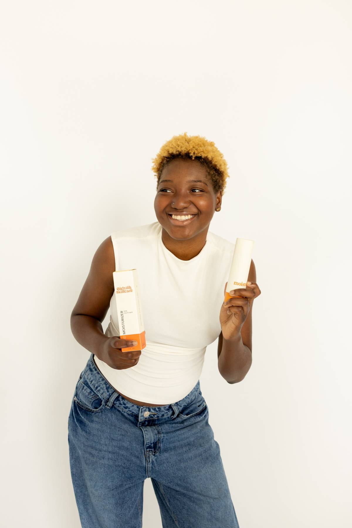 A smiling teen with short curly blonde hair holds a Girlyish Skincare moisturizer bottle in one hand and its packaging box in the other. They are wearing a white sleeveless top and jeans, standing against a plain white background.