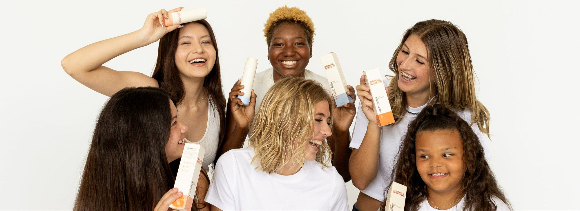 Six smiling girls wearing white tops and jeans holding Girlyish Skincare bottles and boxes, posing together against a white background